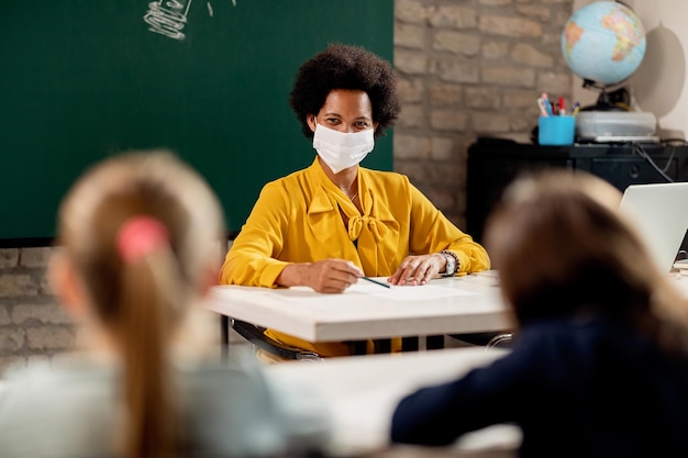 Felice insegnante elementare afroamericana che indossa una maschera facciale mentre tiene una lezione ai suoi studenti in classe.