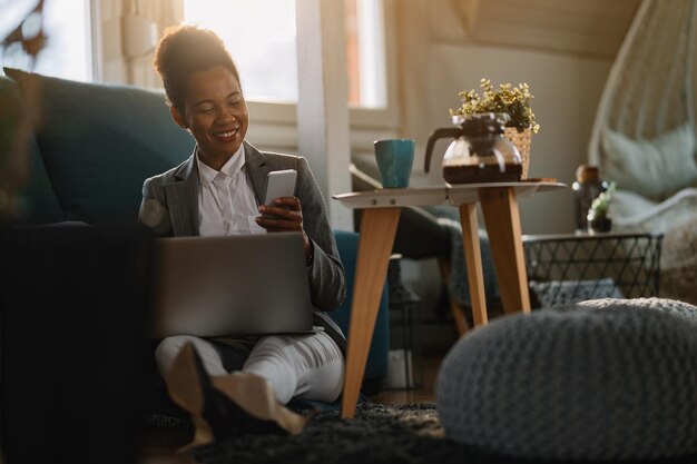 Happy African American entrepreneur reading text message on cell phone while working on laptop at home