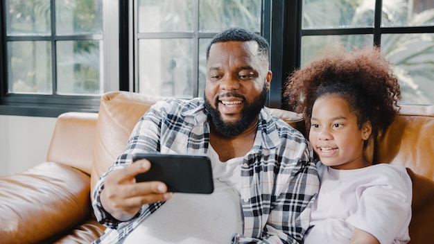 Happy african american family dad and daughter having fun and using mobile phone video call on sofa at house.