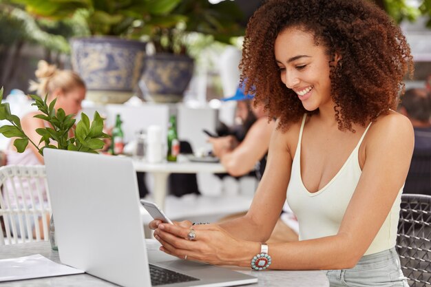 Счастливая афро-американская женщина проверяет банковский баланс на мобильном телефоне, рада получать зарплату, работает внештатно на портативном компьютере в ресторане.