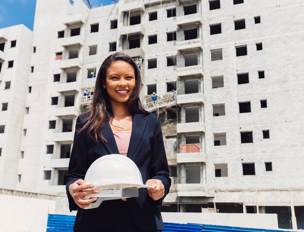 建設中の建物の近くの安全ヘルメットを保持している幸せなアフリカ系アメリカ人女性