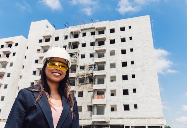 Happy African American lady in safety helmet and eyeglasses near building under construction