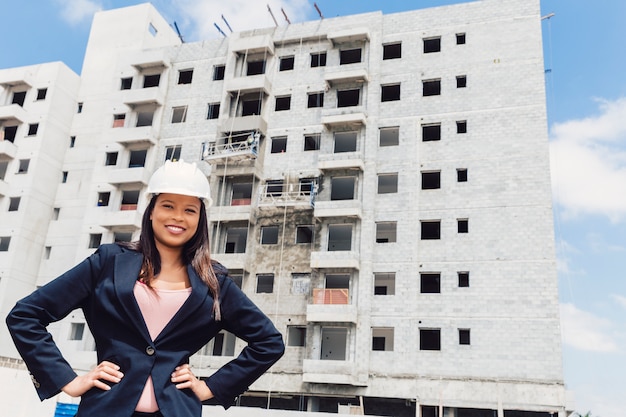 Happy African American lady in safety helmet with hands on hip near building under construction