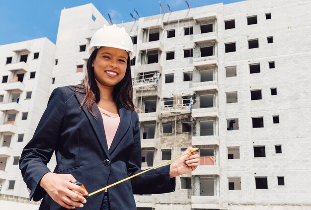 Happy African American lady in safety helmet with measuring tape near building under construction