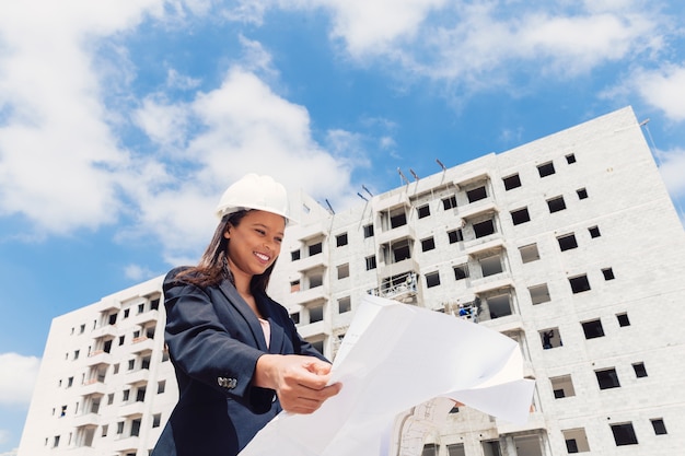 Happy African American lady in safety helmet with paper plan near building under construction
