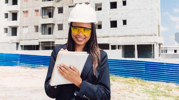 Happy African American lady in safety helmet writing in notebook near building under construction