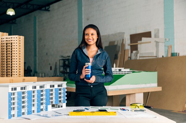 Happy African-American lady with thermos standing near model of building