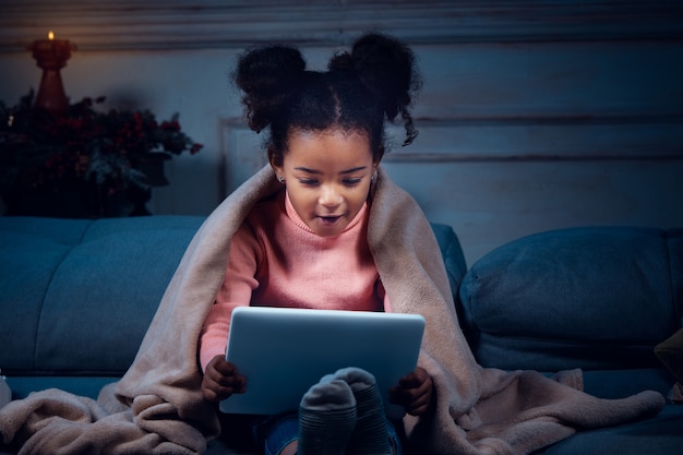 Happy african-american little girl during video call with laptop and home devices, looks delighted