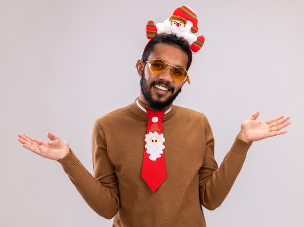 Happy african american man in brown sweater and santa rim on head with funny red tie holding candy cane looking at camera smiling spreading arms to the sides standing over white background