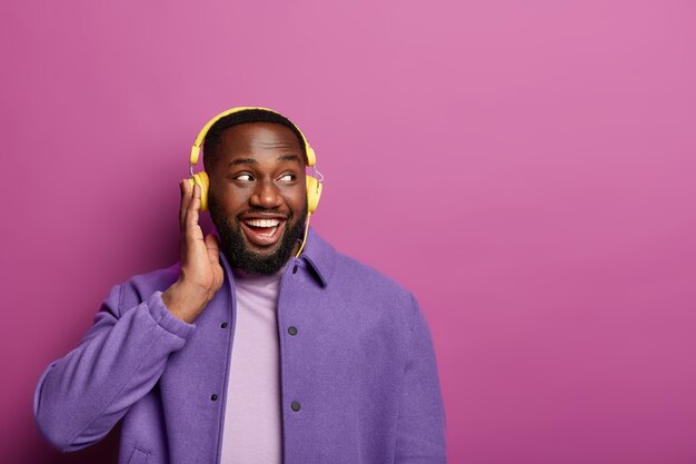Счастливый афроамериканец наслаждается песней в наушниках, слушает приятную мелодию, смотрит в сторону, у него хорошее настроение, носит фиолетовый пиджак