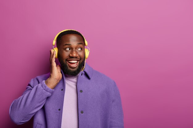 Happy African American man enjoys song playing in headphones, listens pleasant melody, looks aside, has glad mood, wears purple jacket