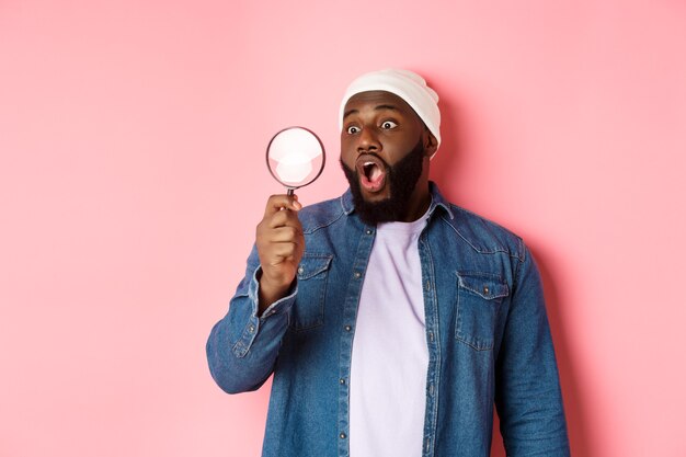 Happy african-american man looking through magnifying glass, smiling amazed, standing against pink background.