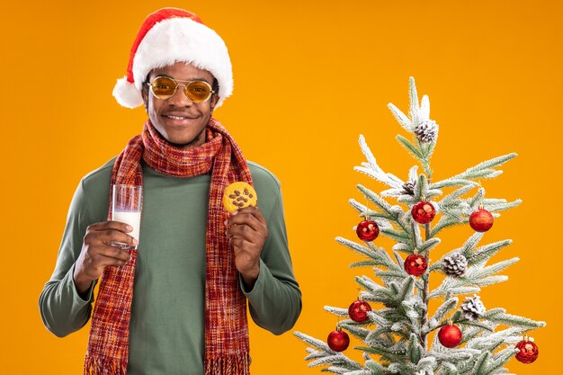 Happy african american man in santa hat and scarf around neck holding glass of milk and cookie smiling standing next to a christmas tree over orange background