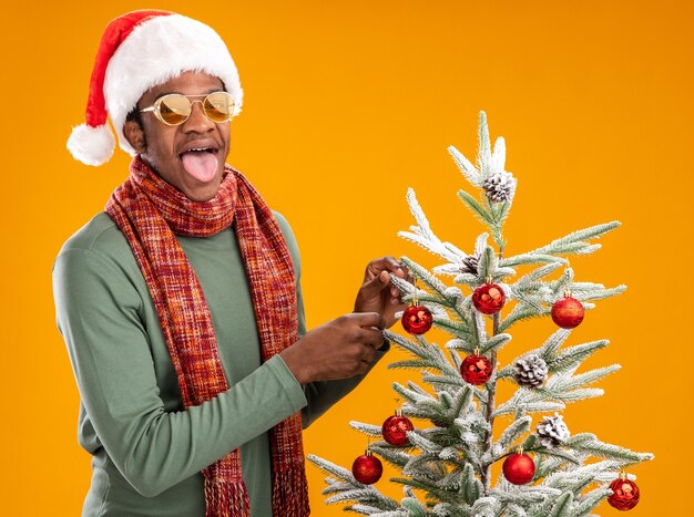 오렌지 배경 위에 크리스마스 트리 옆에 서 혀를 튀어 나와 카메라를보고 목 주위에 산타 모자와 스카프에 행복 아프리카 계 미국인 남자