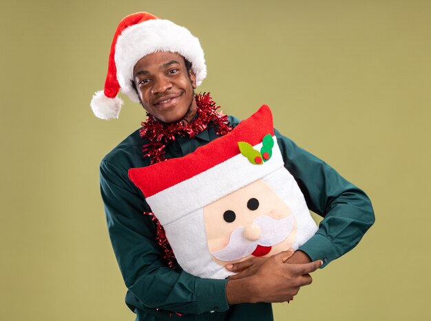 Free photo happy african american man in santa hat with garland holding christmas pillow looking at camera with smile on face standing over green background