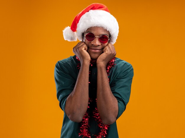 Happy african american man in santa hat with garland wearing glasses looking at camera smiling cheerfully standing over orange background