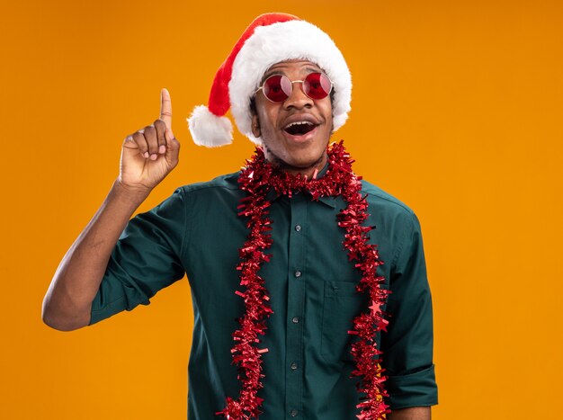 Счастливый афро-американский мужчина в шляпе санта-клауса с гирляндой в очках показывает указательный палец, имеющий новую идею, стоящий над оранжевой стеной
