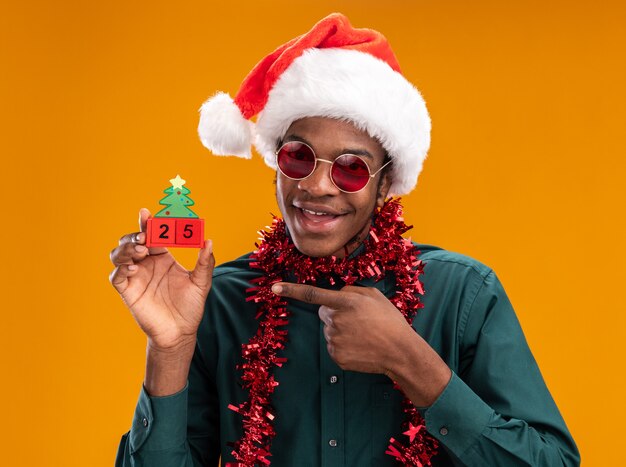 Happy african american man in santa hat with garland wearing sunglasses holding toy cubes with date twenty five pointing with index finger at it standing over orange wall