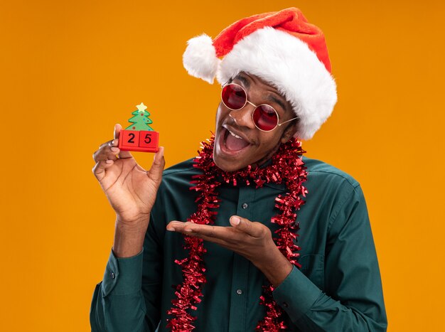 오렌지 배경 위에 팔 서와 함께 제시 날짜 25 장난감 큐브를 들고 선글라스를 착용하는 갈 랜드와 산타 모자에 행복 한 아프리카 계 미국인 남자
