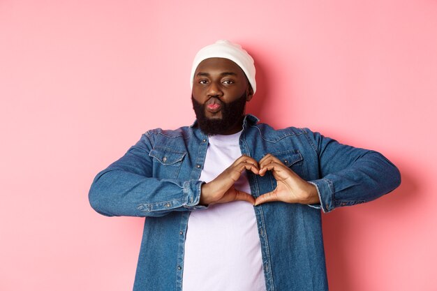 Happy african-american man showing heart sign, I love you gesture, pucker lips for kiss while standing over pink background