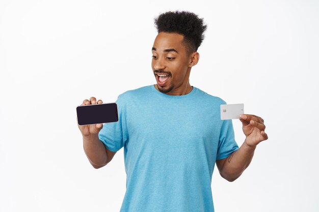 Счастливый афроамериканец показывает приложение для покупок, горизонтальный экран смартфона и кредитную карту, улыбается, рекомендует приложение, стоя на белом фоне.