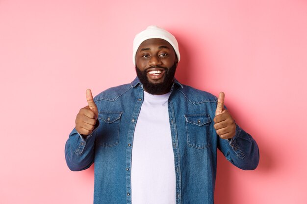 Happy african-american man showing thumbs-up in approval, like something good, praising great deal, standing over pink background