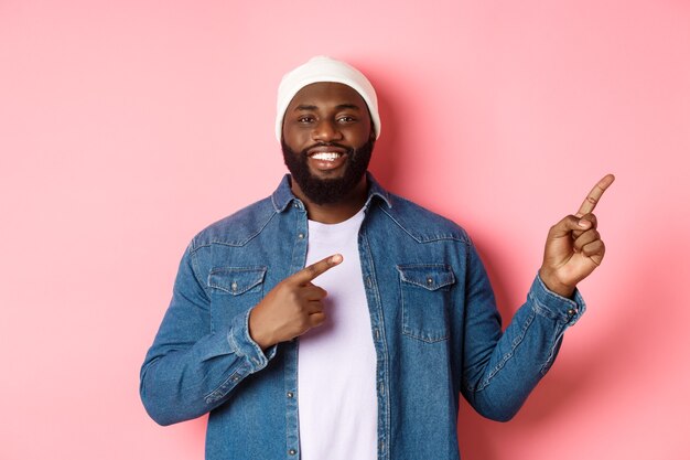 Happy african-american man smiling, pointing fingers right and showing promo, making announcement, standing over pink background.