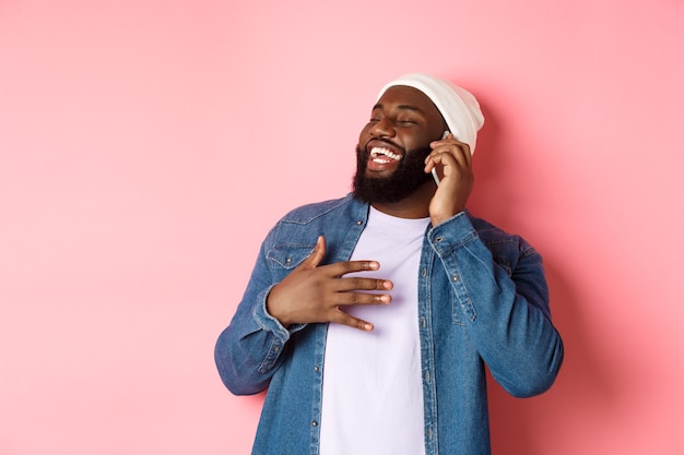 Счастливый афро-американский мужчина разговаривает по мобильному телефону, смеется и улыбается, стоя в шапке и джинсовой рубашке на розовом фоне