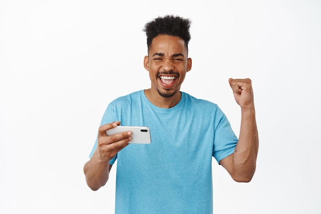 幸せなアフリカ系アメリカ人の男が携帯電話のビデオゲームで勝ち、唱え、祝い、喜んで悲鳴を上げ、笑顔で、白のスマートフォンアプリを使用します