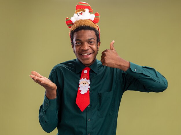 Happy african american man with funny santa rim and red tie looking at camera showing thumbs up and down standing over green background