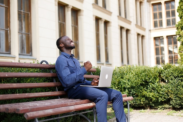 Happy African American man works on his laptop sitting on the bench outside