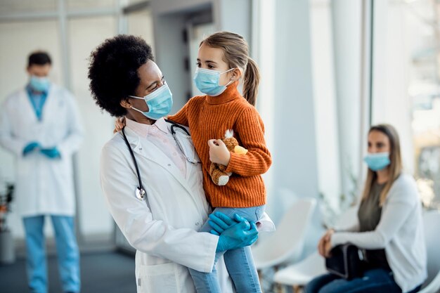 Happy African American pediatrician holding a small girl in the hallway at medical clinic