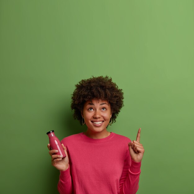 Happy African American woman drinks fresh fruit smoothie, holds beverage made of super foods in glass bottle, points index finger above on green wall, being in good mood. Healthy lifestyle