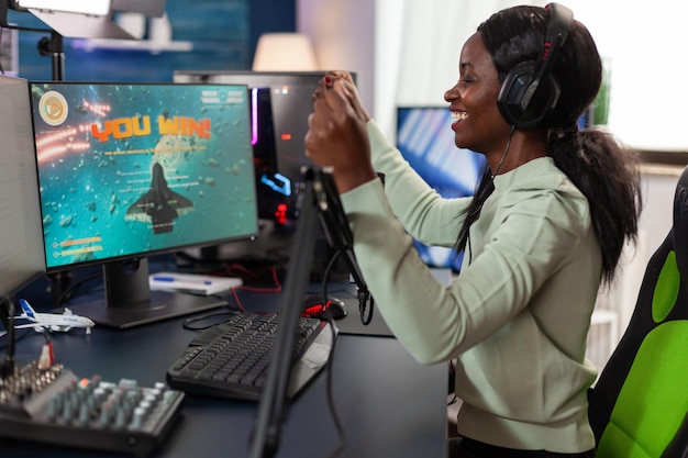 Foto gratuita giocatore felice della donna afroamericana che indossa l'auricolare vincendo la competizione di videogiochi che parla nel microfono con i giocatori. placcatore eccitato che gioca a giochi sparatutto spaziali utilizzando un computer rgb. diretta streaming