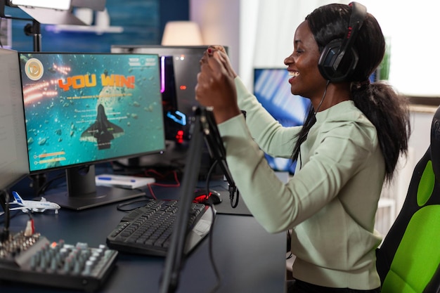 Счастливая африканская американка-геймер в гарнитуре, выигравшая соревнование по видеоиграм, разговаривает в микрофон с игроками. Возбужденный игрок, играющий в космические стрелялки на компьютере с RGB-подсветкой. Прямая трансляция