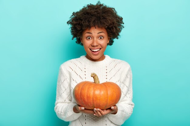 Happy African American woman holds orange pumpkin, wears white sweater,  smiles gently, isolated on blue wall