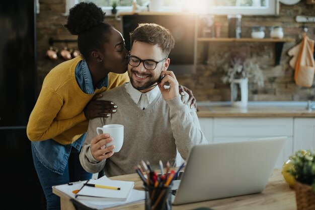 Счастливая афроамериканка целует своего мужа, который работает дома на ноутбуке