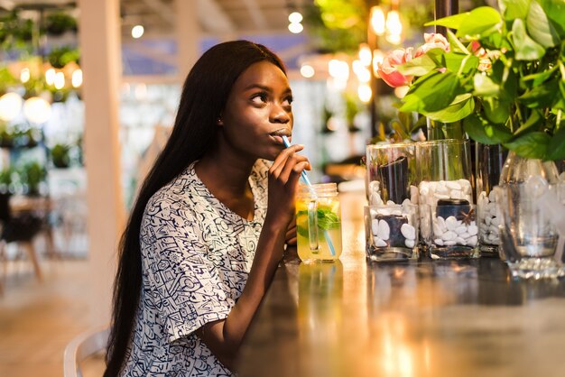 Happy African-American woman with glass of natural lemonade in cafe. Detox drink