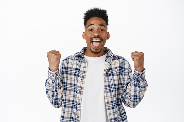 Happy african american young man celebrating, winning, rooting for team, clench and make fist pump to triumph win, achieve goal, looking amazed at camera, white background.