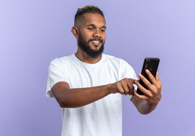 Happy african american young man in white t-shirt holding smartphone looking at it writing message smilign cheerfully