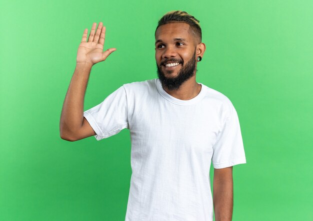Счастливый афро-американский молодой человек в белой футболке смотрит в сторону, весело улыбаясь, машет рукой