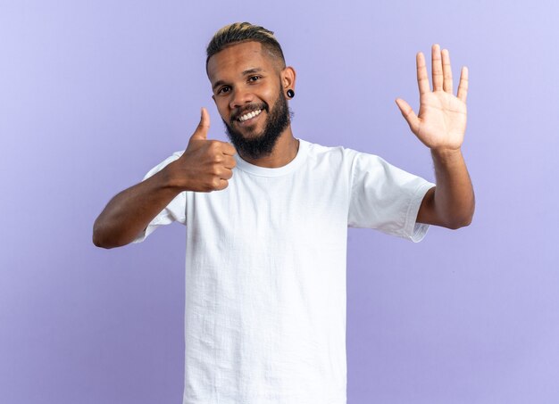 유쾌 하 게 웃 고 엄지 손가락을 보여주는 손으로 흔들며 카메라를보고 흰색 티셔츠에 행복 한 아프리카 계 미국인 젊은 남자