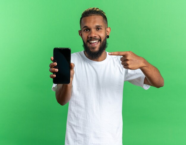 幸せなアフリカ系アメリカ人の若い男が白いTシャツを着て、カメラの笑顔を見て人差し指でスマートフォンを指しています