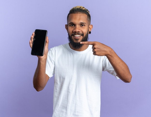Happy african american young man in white t-shirt showing smartphone pointing