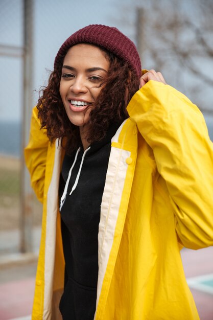 野外を歩いている黄色のコートを着て幸せなアフリカの巻き毛の若い女性