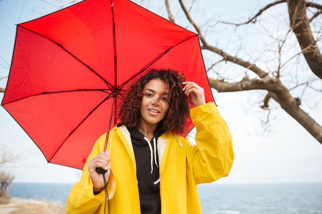 Happy african curly young lady wearing yellow coat with umbrella.