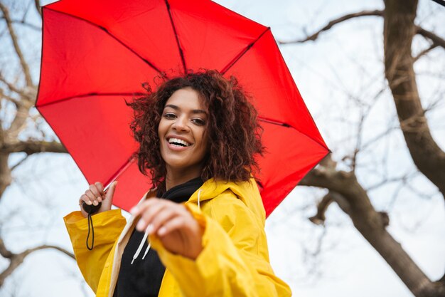 Happy african curly young lady wearing yellow coat with umbrella.