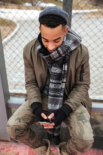 Happy african man sitting and using smartphone outdoors in autumn