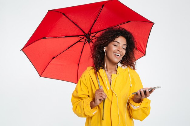 Happy african woman in raincoat hiding under umbrella and listening music on her smartphone over white background
