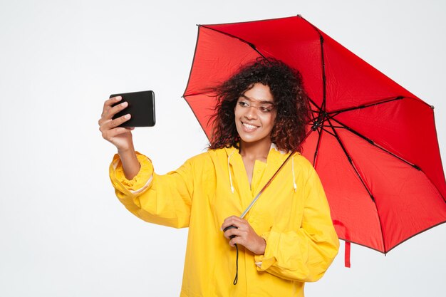 Happy african woman in raincoat hiding under umbrella and making selfie on smartphone over white background
